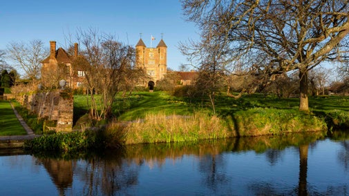 A view from the orchard at Sissinghurst Castle Garden, Kent, across the moat towards the Elizabethan Tower. The bright blue skies and bare trees are reflected in the water.